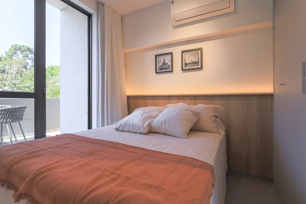 Quarto com cama posta, manta cor de telha, quadros e janela para o jardim.