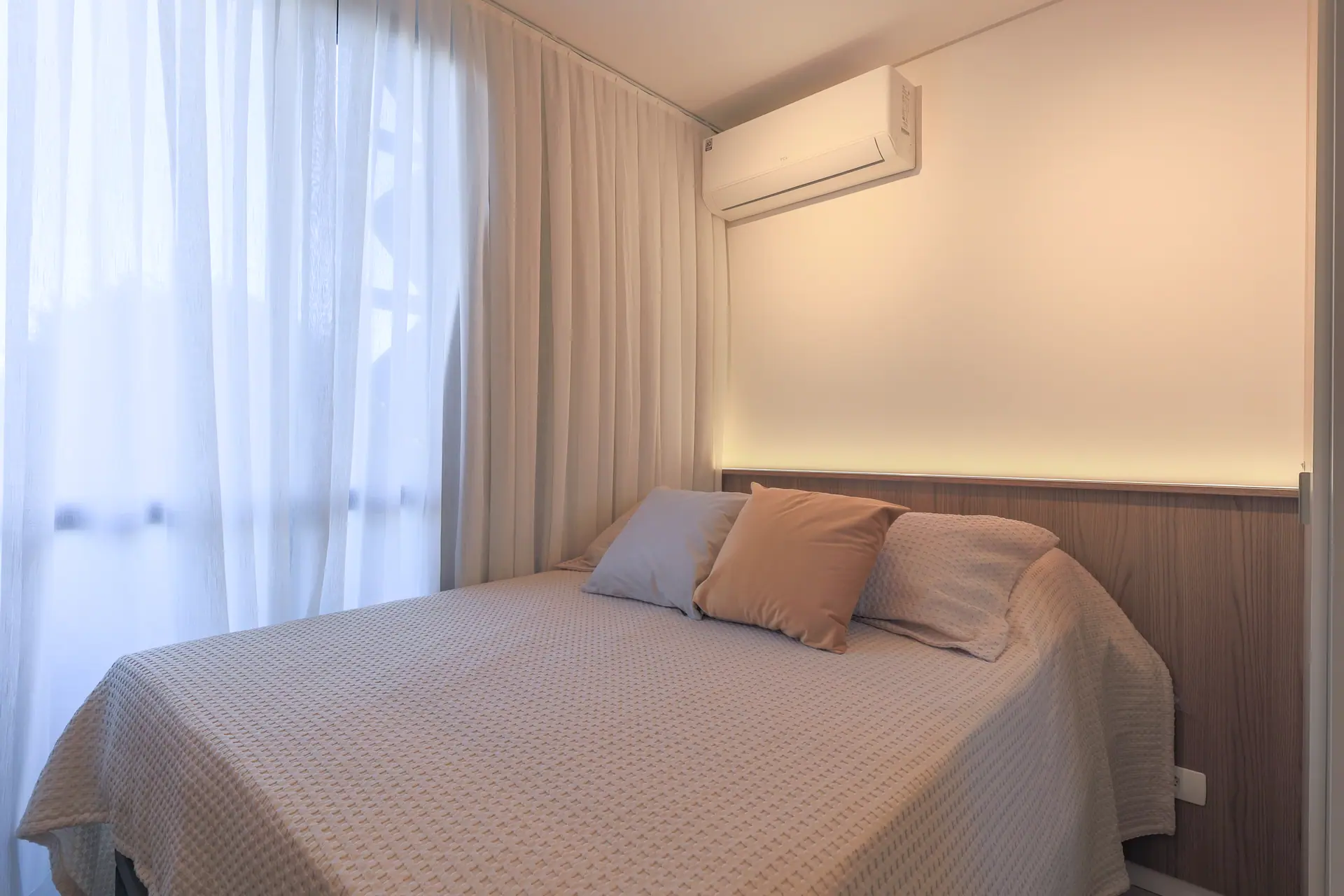 Cama posta com almofadas, cabeceira em madeira e luz embutida na parede.