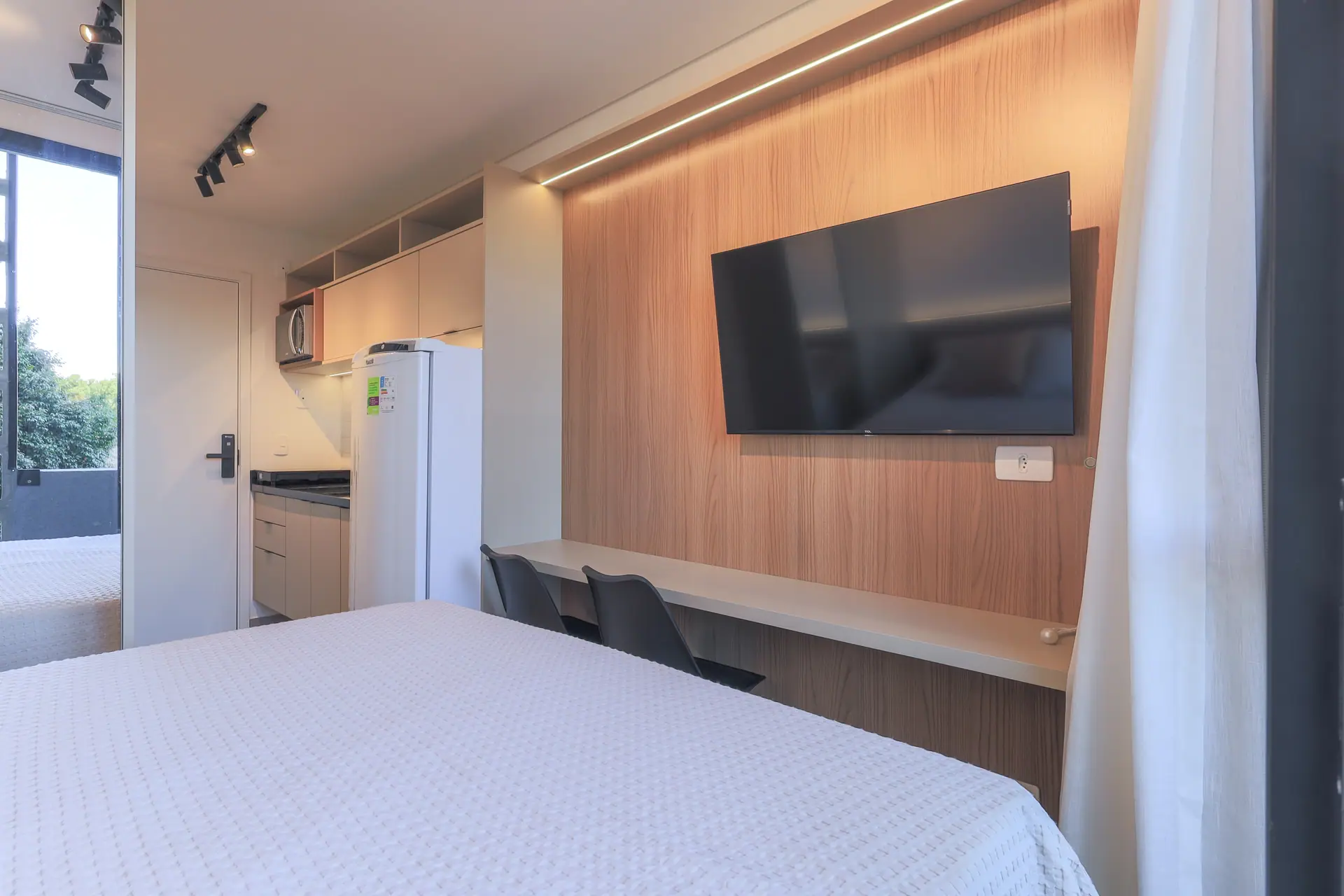 Studio visto da janela: cama, bancada com duas cadeiras, painel de madeira e a cozinha ao fundo.