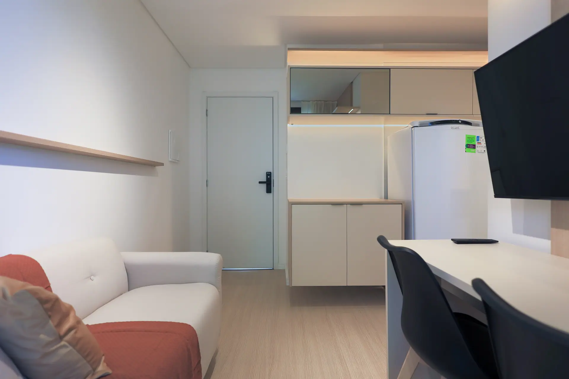 Sala com sofá e bancada, e a cozinha com geladeira e armários ao fundo.