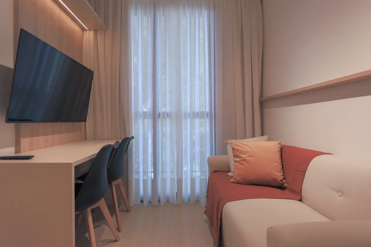 Sala com sofá e almofadas, bancada com cadeiras e painel com TV junto à janela.